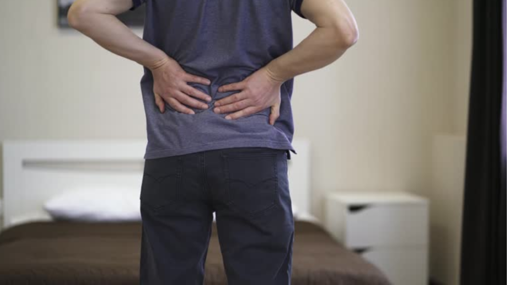 朝起きて、腰の痛みを感じ、腰に手を当てる人物の画像